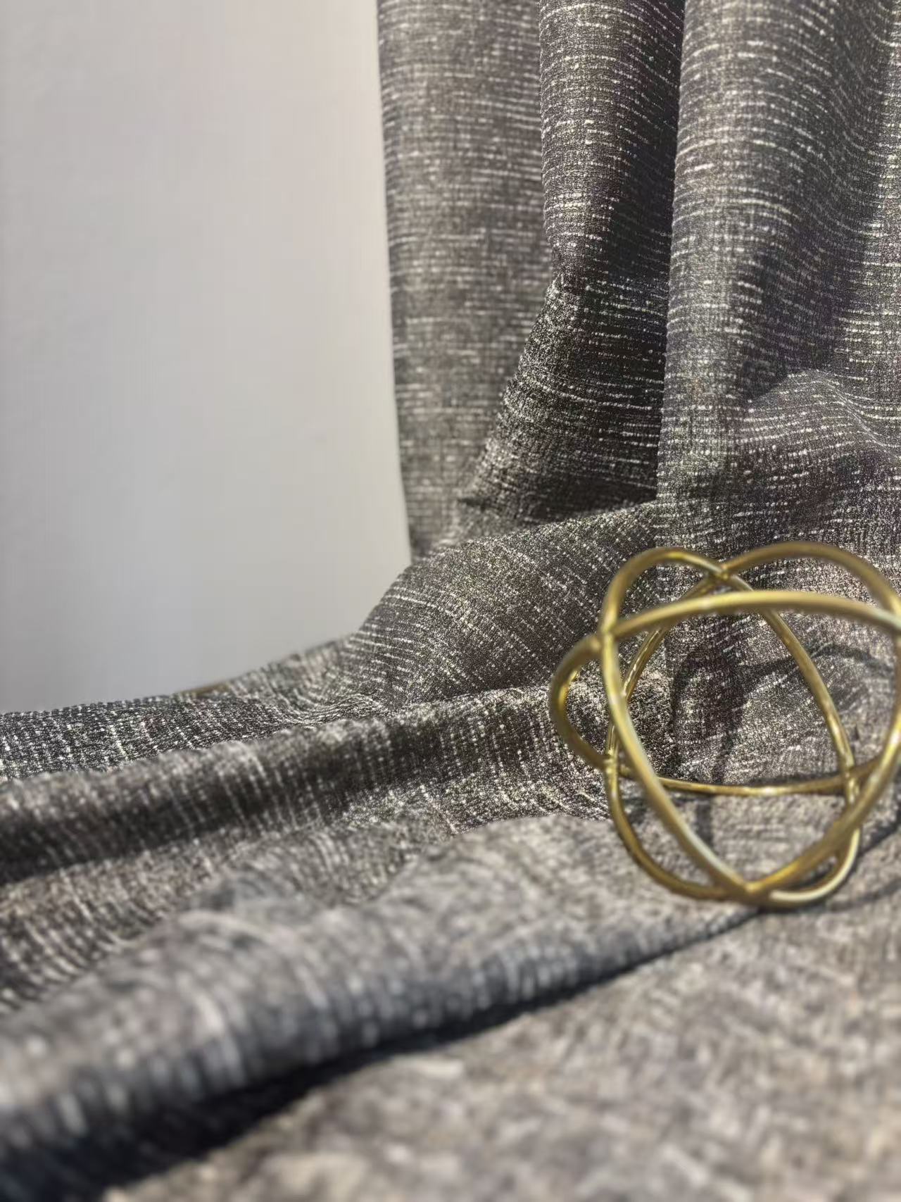 这是一张展示灰色混纺面料的特写图片。布料呈现出细腻的横向纹理，并在光线下隐约透出细微的闪光质感，显得高级且有层次。面料自然垂坠，褶皱柔和，旁边放置了一个金色的几